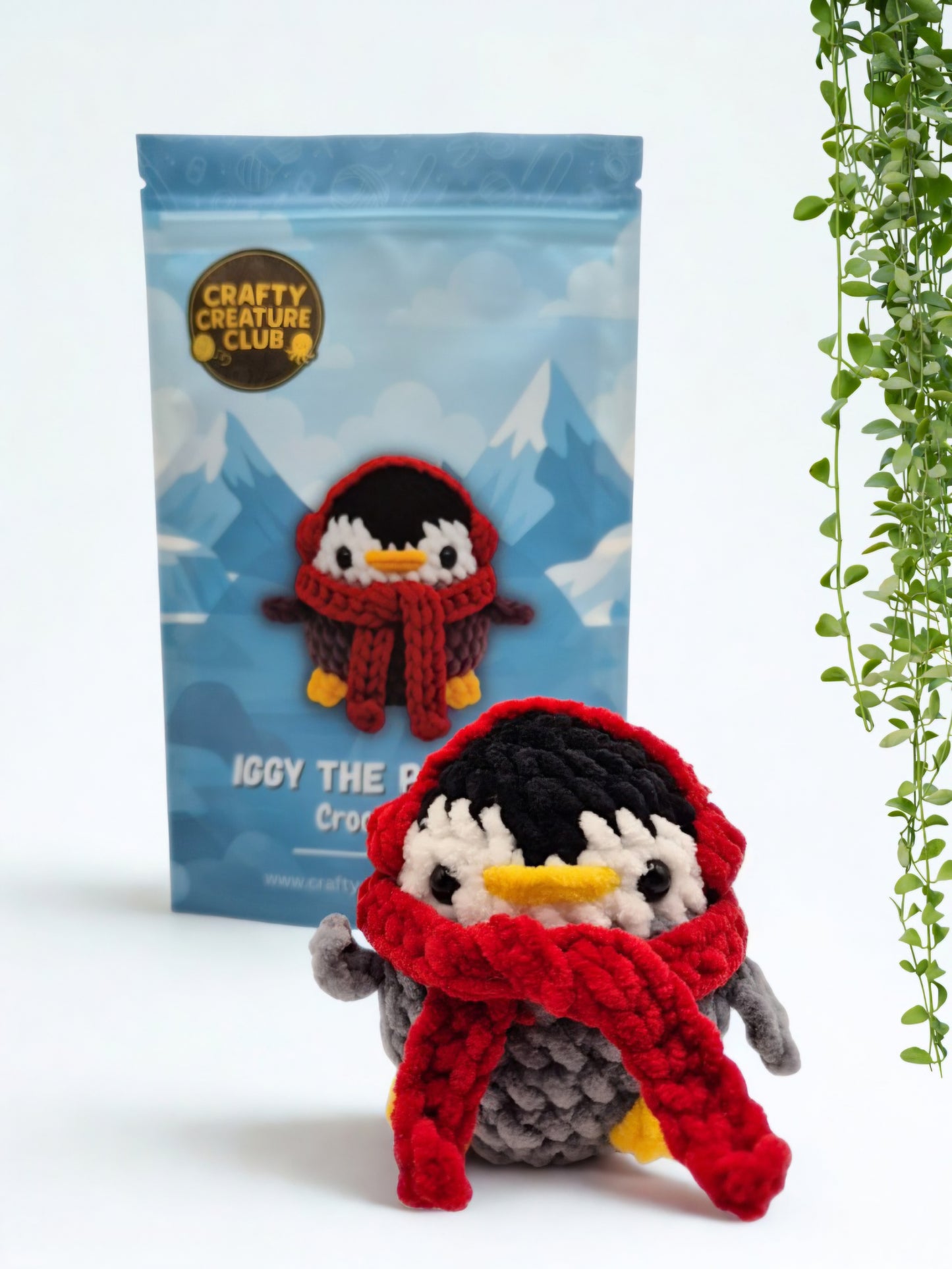 Iggy penguin crochet kit shown with packaging and finished plush penguin wearing a red scarf and earmuffs